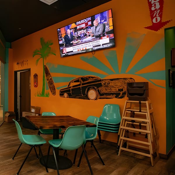 Hacienda Tacos interior with vibrant orange walls, a TV, and a mural of a car and palm tree.