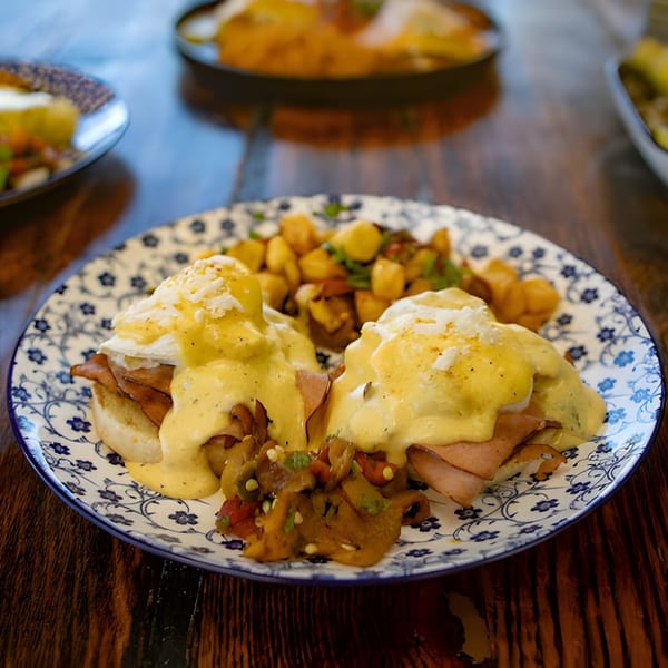 Hacienda Tacos' Eggs Benedict with ham, hollandaise, and roasted potatoes on a patterned plate.