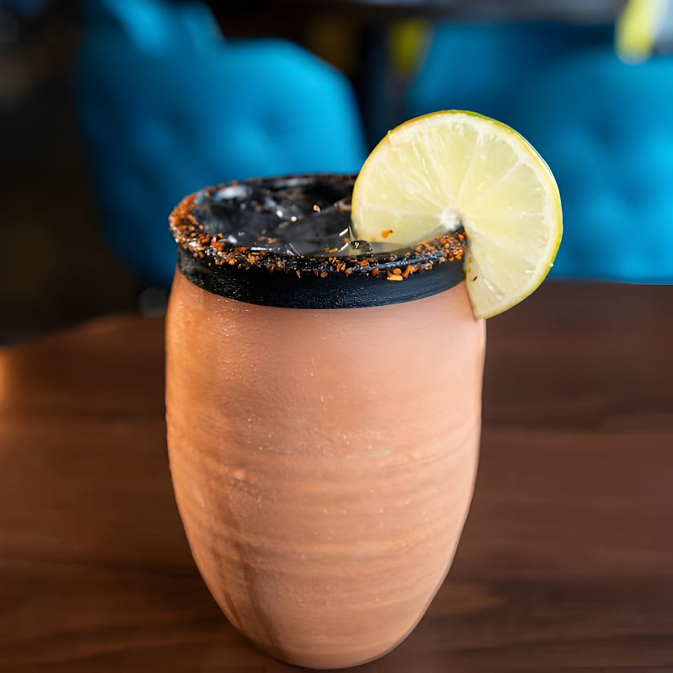 A Hacienda Tacos Mexican drink in a clay cup with a chili-lime rim and a lime wedge.