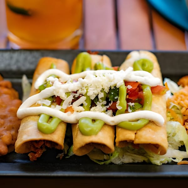 Three Hacienda Tacos taquitos topped with crema, avocado sauce, and crumbled cheese, served with beans and rice.