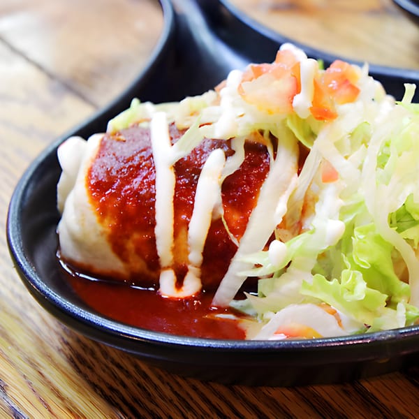 A Hacienda Tacos burrito smothered in red sauce, crema, lettuce, and diced tomatoes in a black dish.